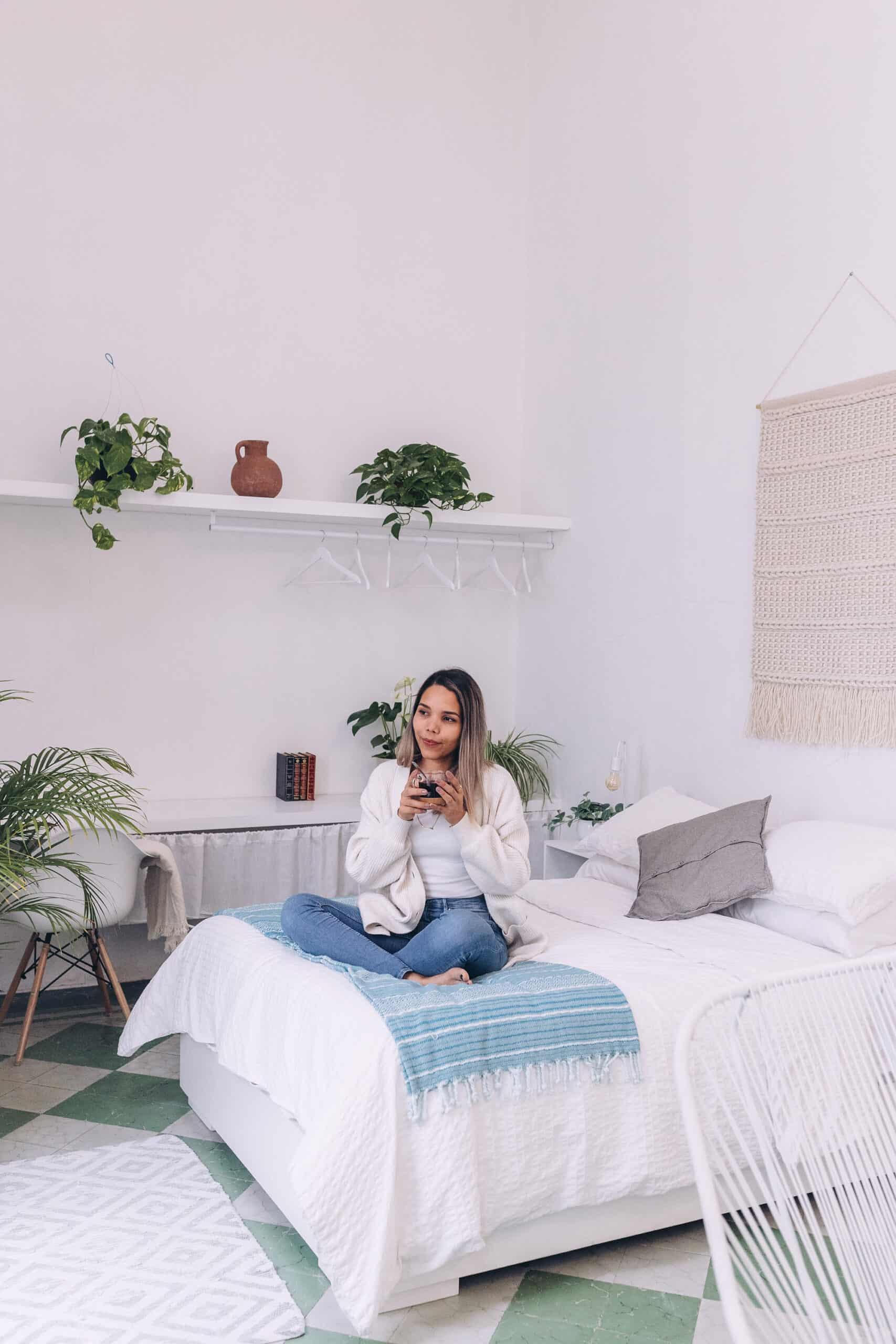 A girl sitting on her bed drinking tea. The bed has white sheets and there are plants on book shelves behind her.