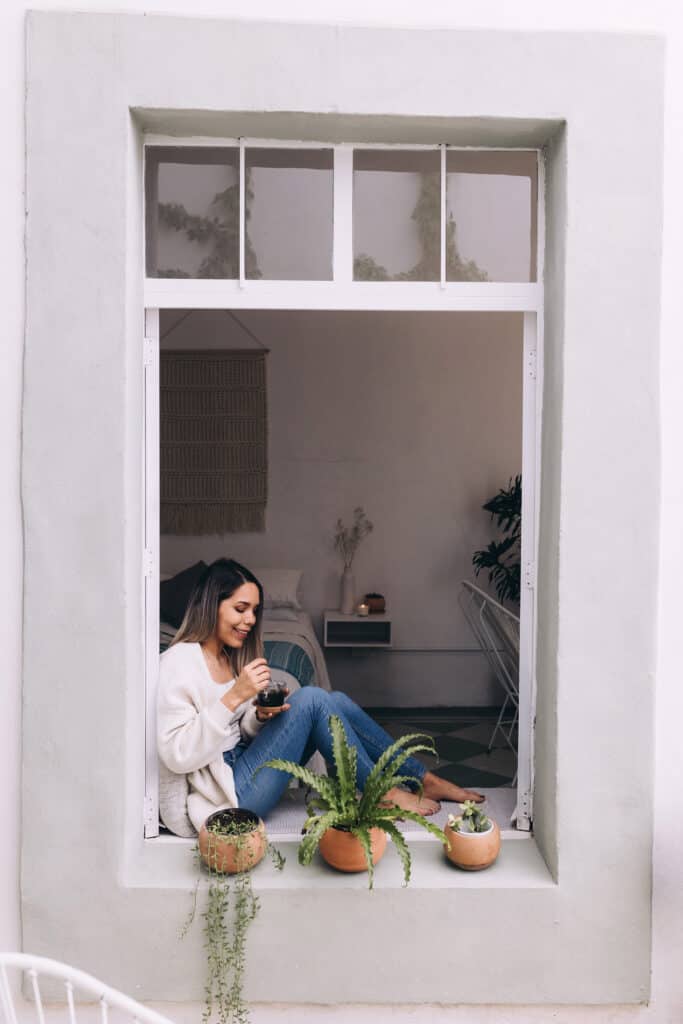 A woman sitting on a windowsill drinking coffee with plants sitting next to her.