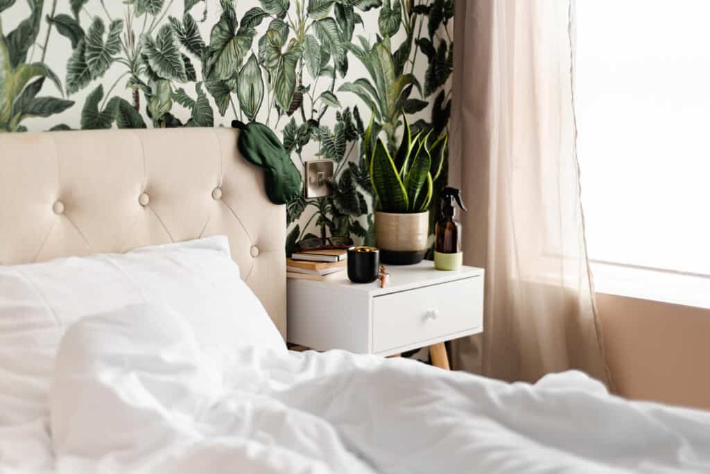 A bed with white sheets by plant leaf wallpaper and a plant on the nightstand. 